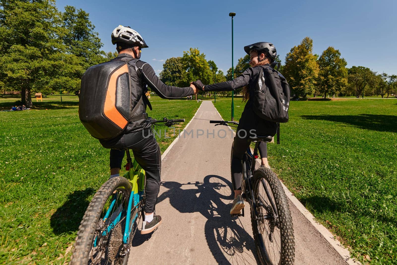 A sweet couple, adorned in cycling gear, rides their bicycles, their hands interlocked in a romantic embrace, capturing the essence of love, adventure, and joy on a sunlit path.