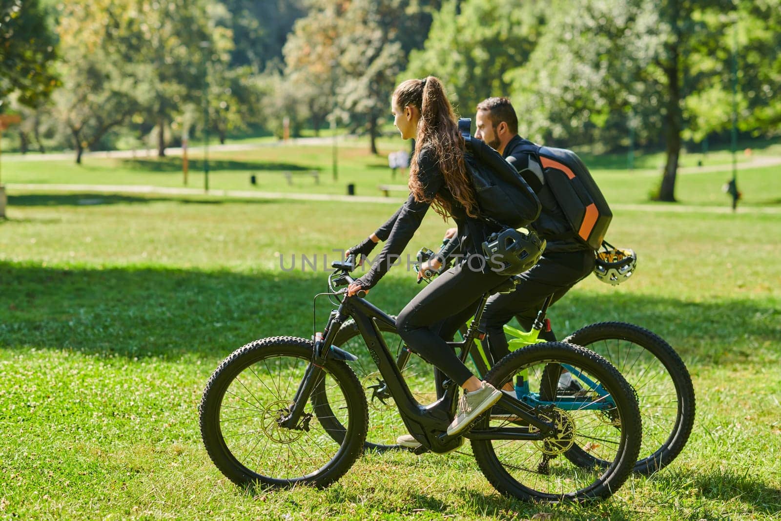 A blissful couple, adorned in professional cycling gear, enjoys a romantic bicycle ride through a park, surrounded by modern natural attractions, radiating love and happiness.