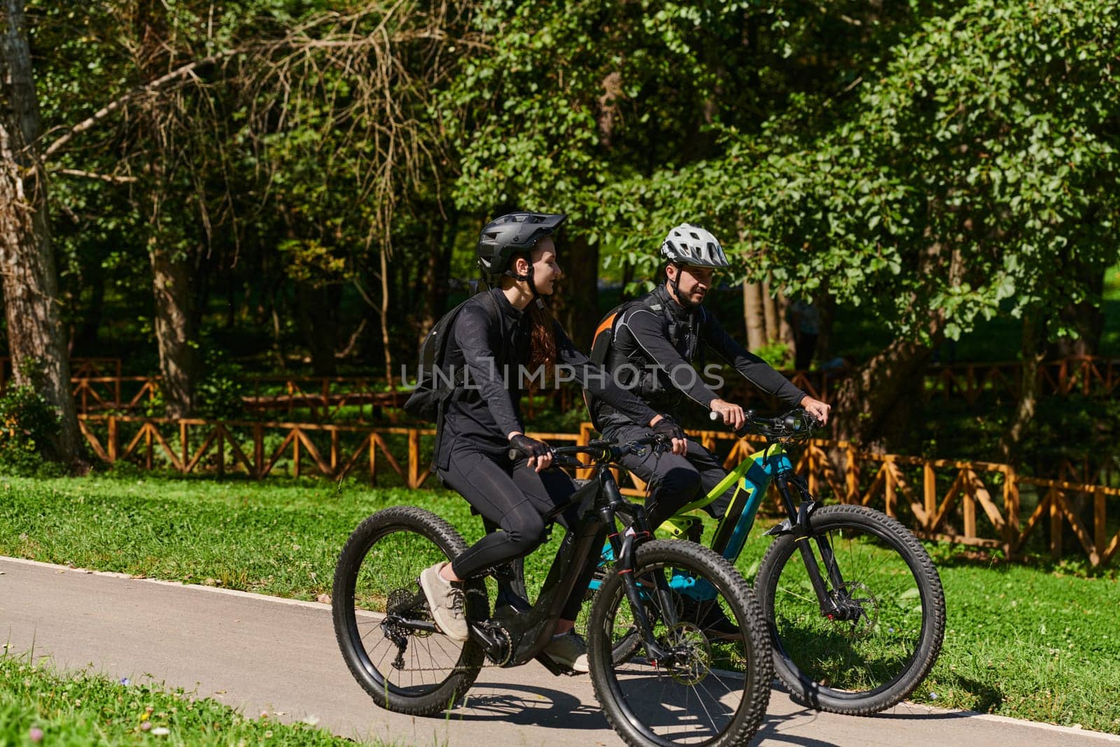 A blissful couple, adorned in professional cycling gear, enjoys a romantic bicycle ride through a park, surrounded by modern natural attractions, radiating love and happiness by dotshock