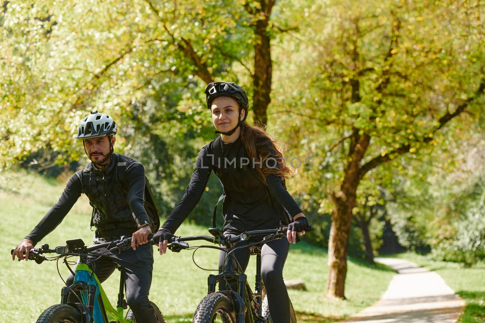 A blissful couple, adorned in professional cycling gear, enjoys a romantic bicycle ride through a park, surrounded by modern natural attractions, radiating love and happiness by dotshock