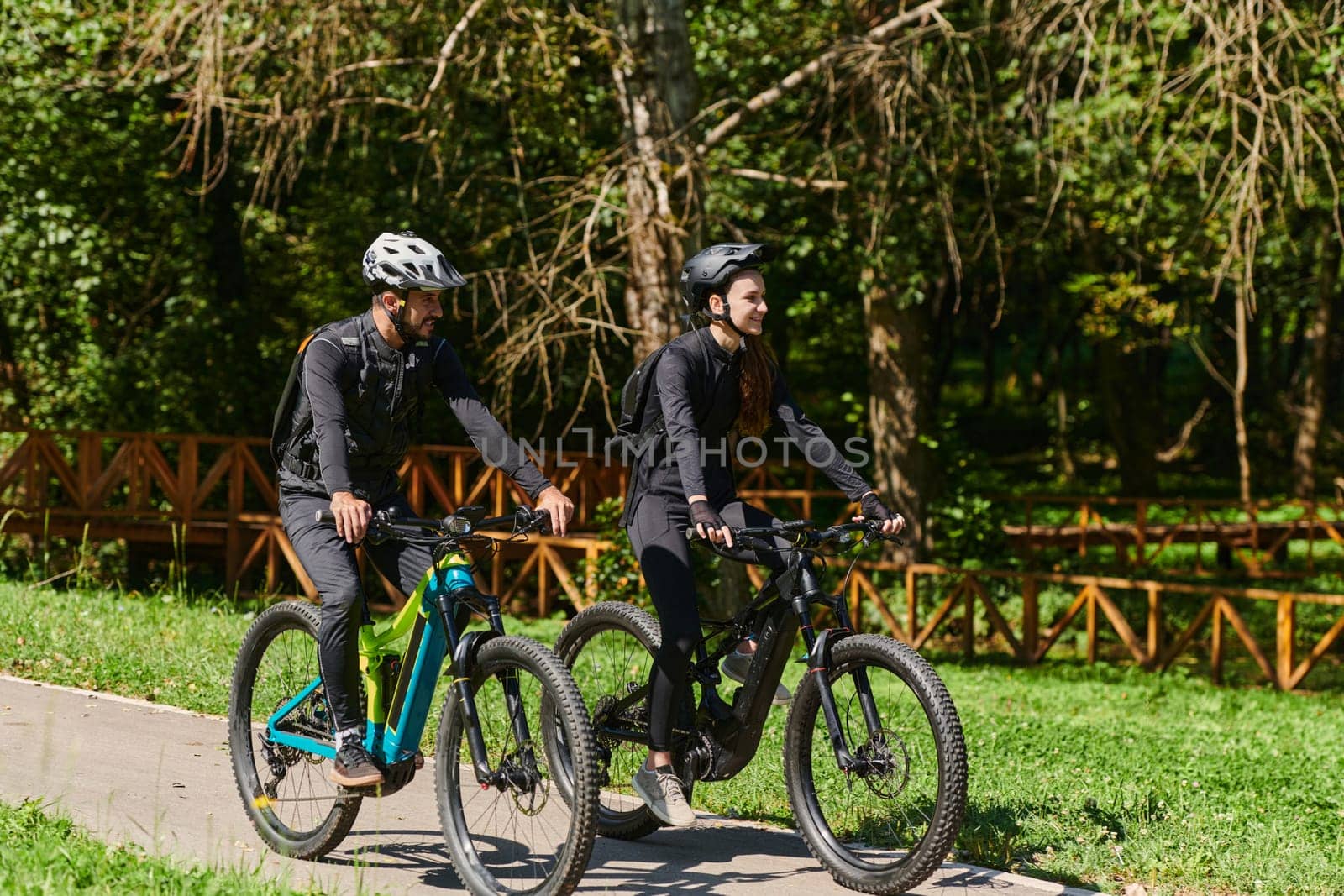 A blissful couple, adorned in professional cycling gear, enjoys a romantic bicycle ride through a park, surrounded by modern natural attractions, radiating love and happiness by dotshock