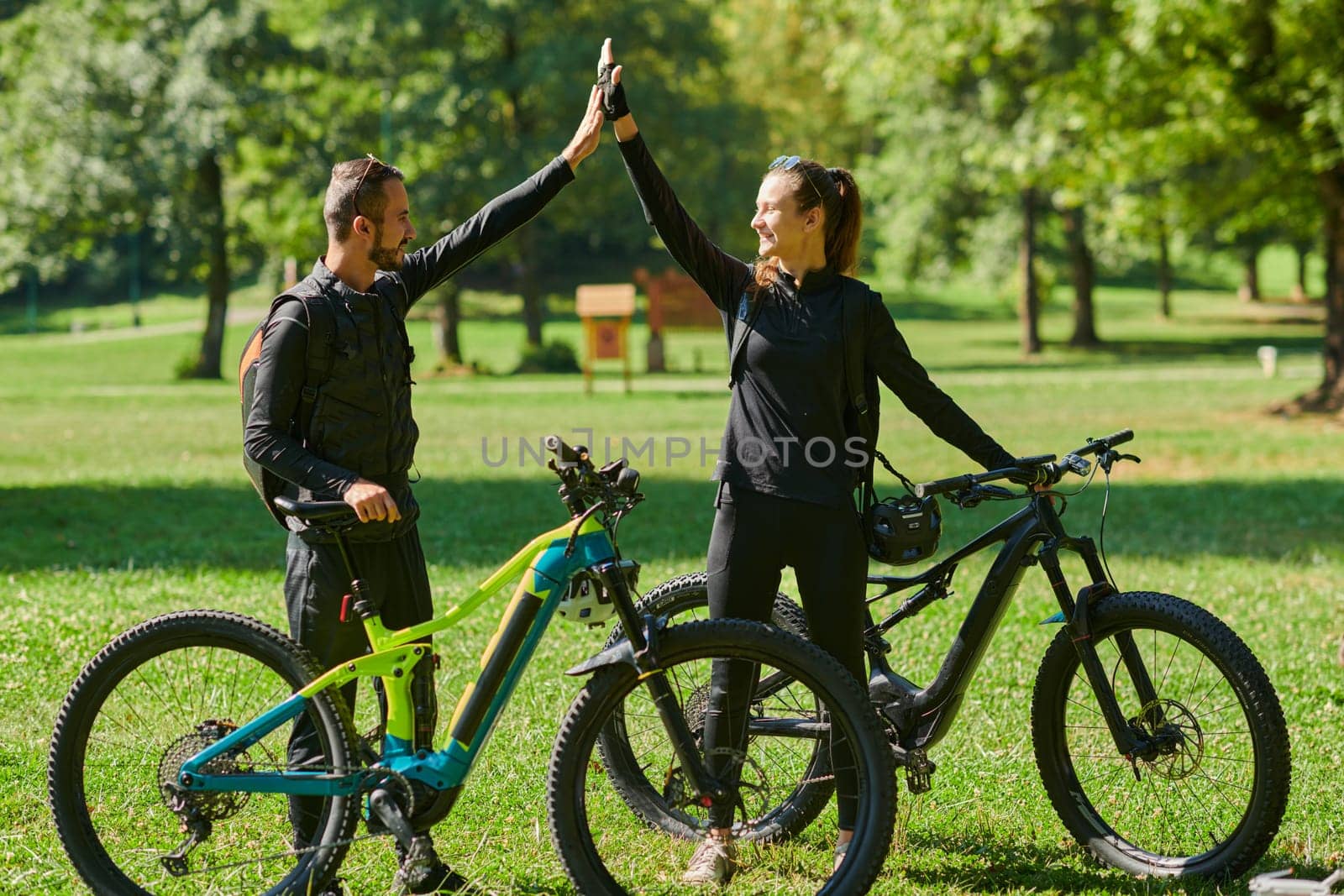 A sweet couple, adorned in cycling gear, rides their bicycles, their hands interlocked in a romantic embrace, capturing the essence of love, adventure, and joy on a sunlit path.