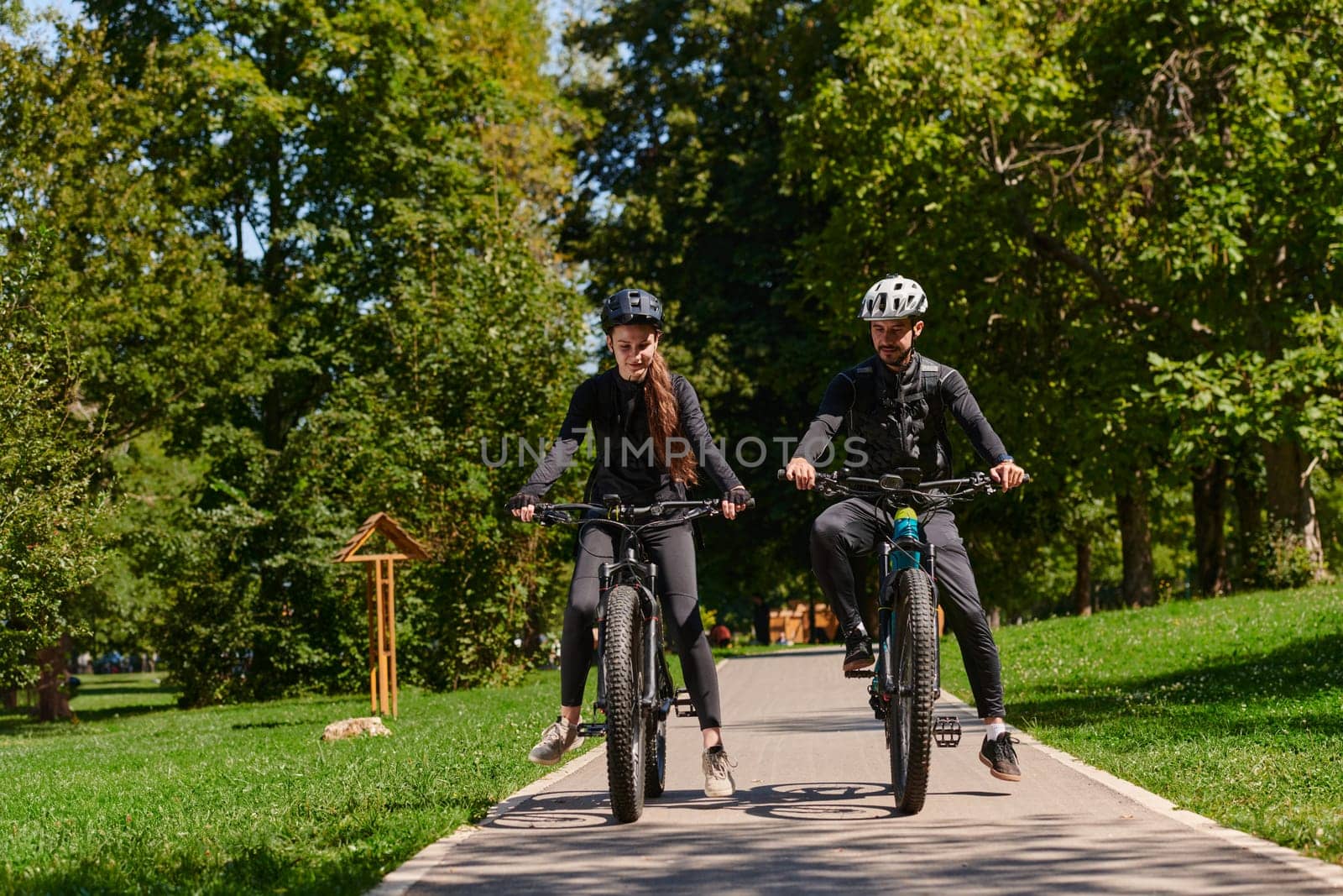 A blissful couple, adorned in professional cycling gear, enjoys a romantic bicycle ride through a park, surrounded by modern natural attractions, radiating love and happiness by dotshock