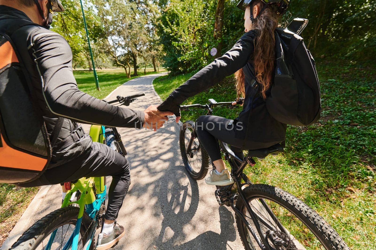 A sweet couple, adorned in cycling gear, rides their bicycles, their hands interlocked in a romantic embrace, capturing the essence of love, adventure, and joy on a sunlit path.