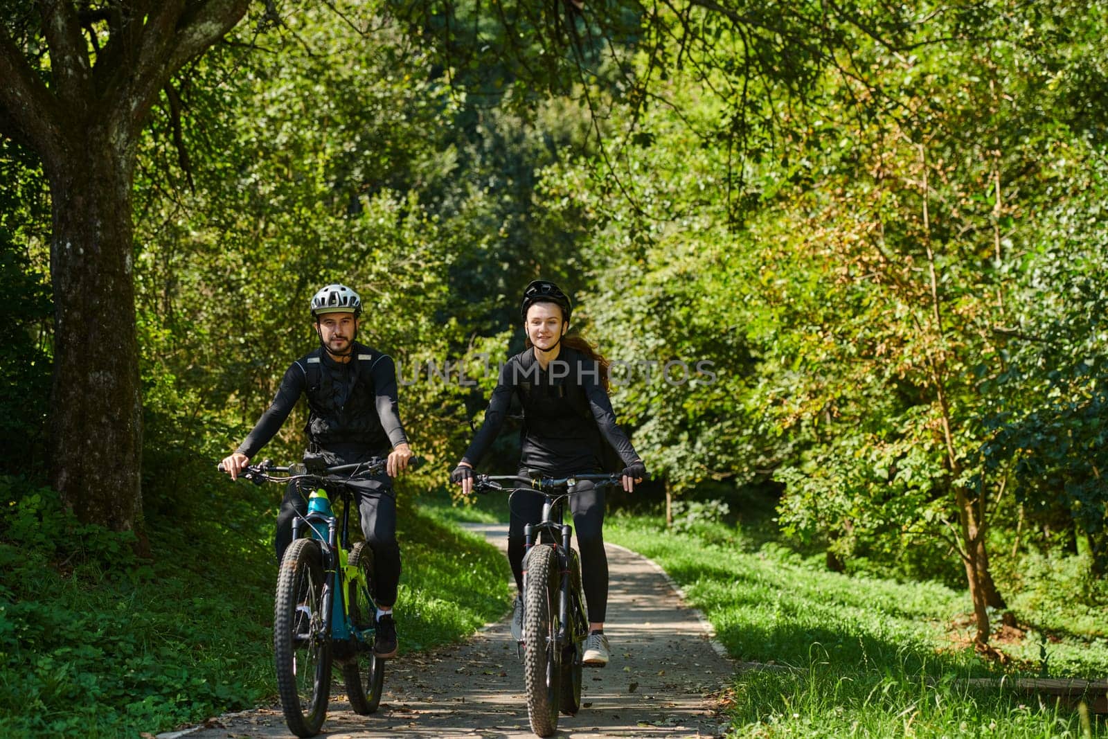 A blissful couple, adorned in professional cycling gear, enjoys a romantic bicycle ride through a park, surrounded by modern natural attractions, radiating love and happiness by dotshock