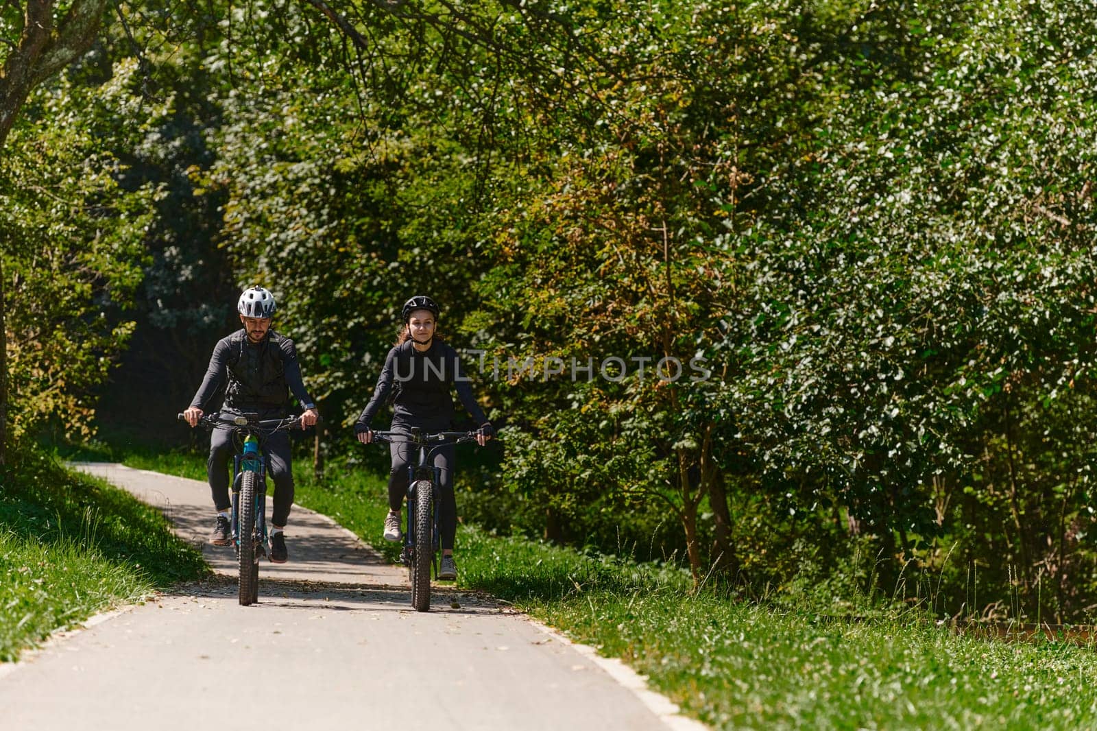 A blissful couple, adorned in professional cycling gear, enjoys a romantic bicycle ride through a park, surrounded by modern natural attractions, radiating love and happiness by dotshock