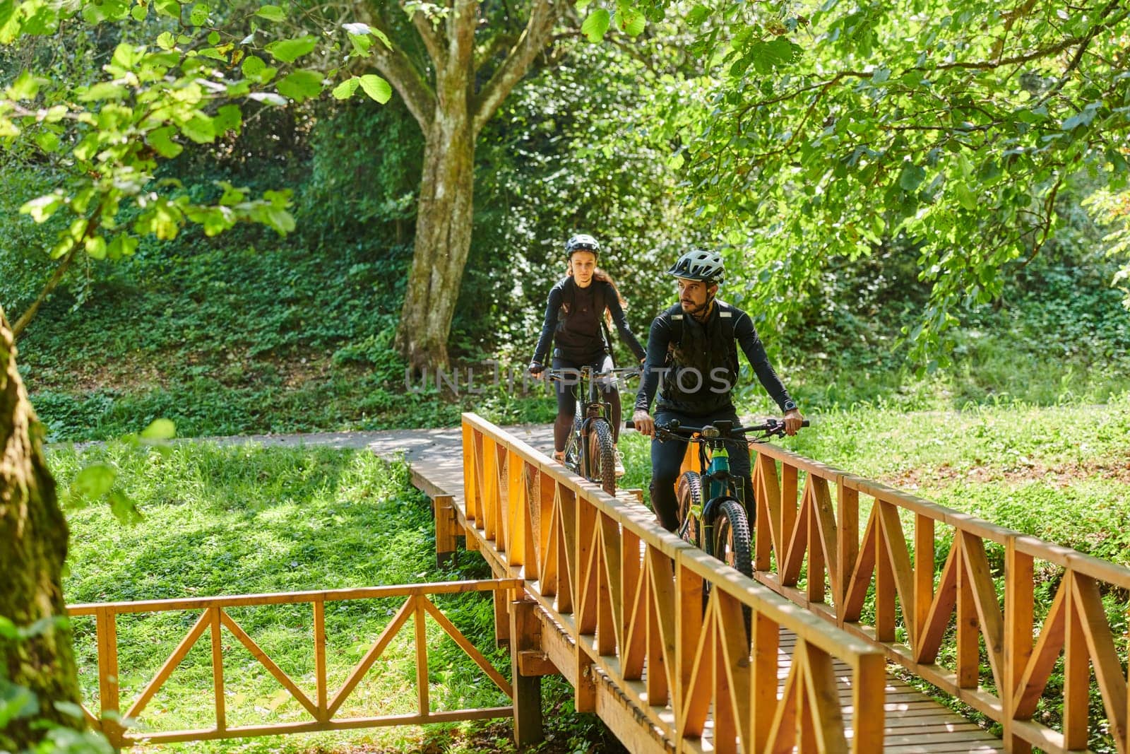 A blissful couple, adorned in professional cycling gear, enjoys a romantic bicycle ride through a park, surrounded by modern natural attractions, radiating love and happiness.