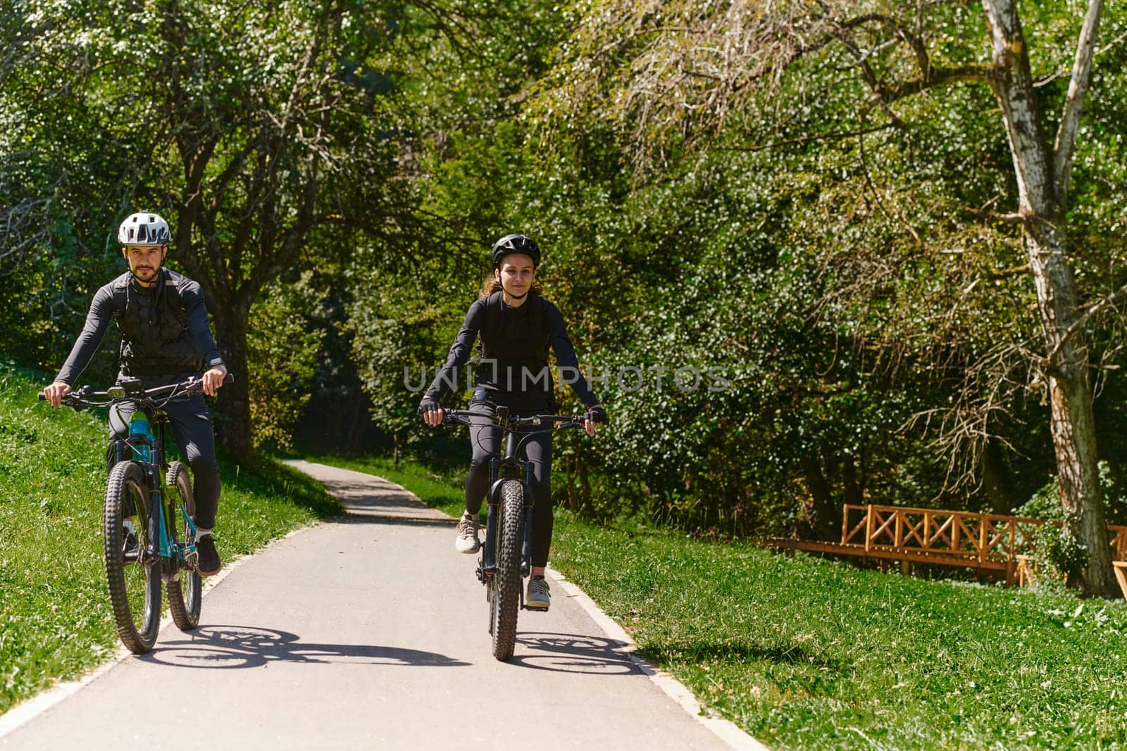 A blissful couple, adorned in professional cycling gear, enjoys a romantic bicycle ride through a park, surrounded by modern natural attractions, radiating love and happiness.