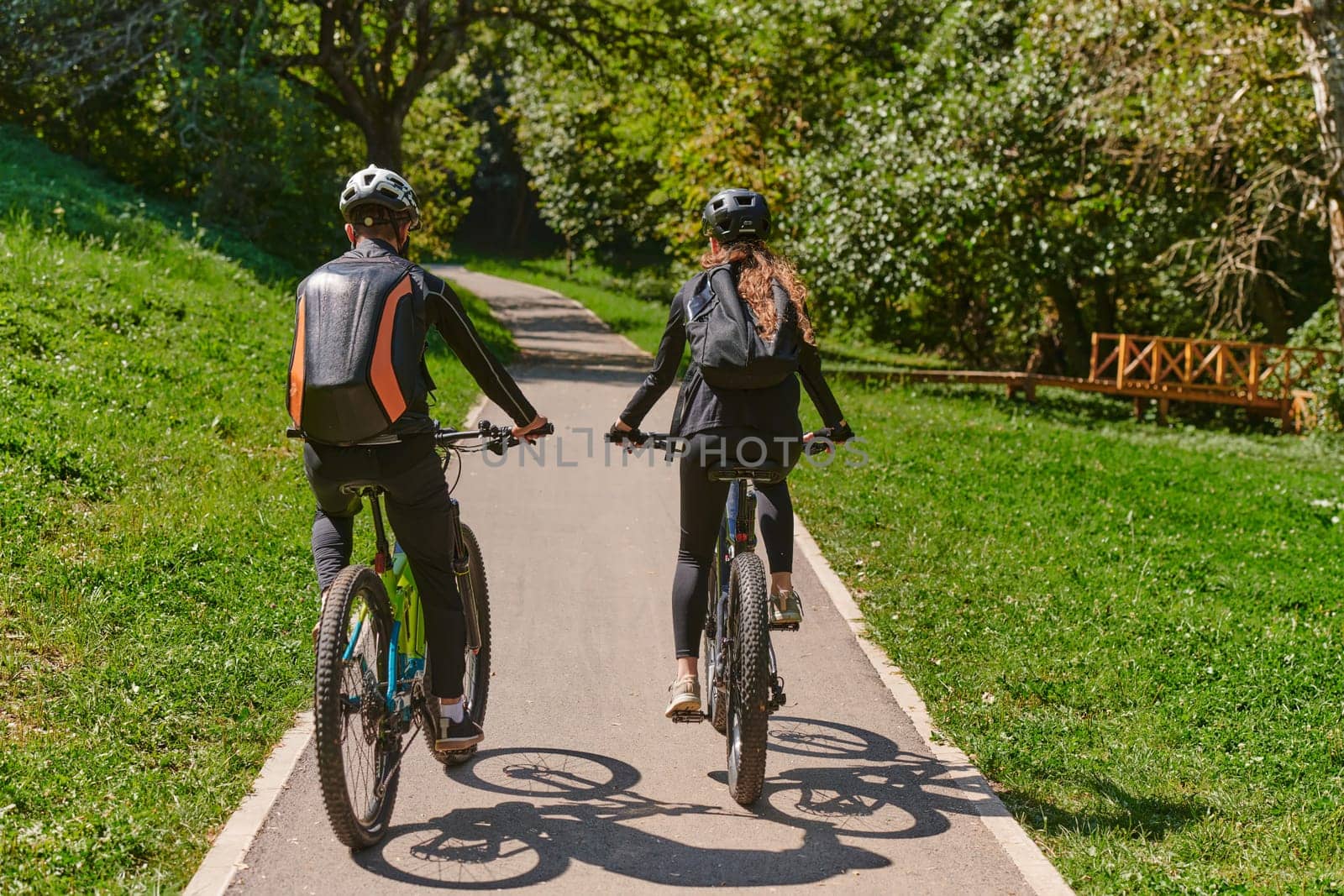 A blissful couple, adorned in professional cycling gear, enjoys a romantic bicycle ride through a park, surrounded by modern natural attractions, radiating love and happiness by dotshock