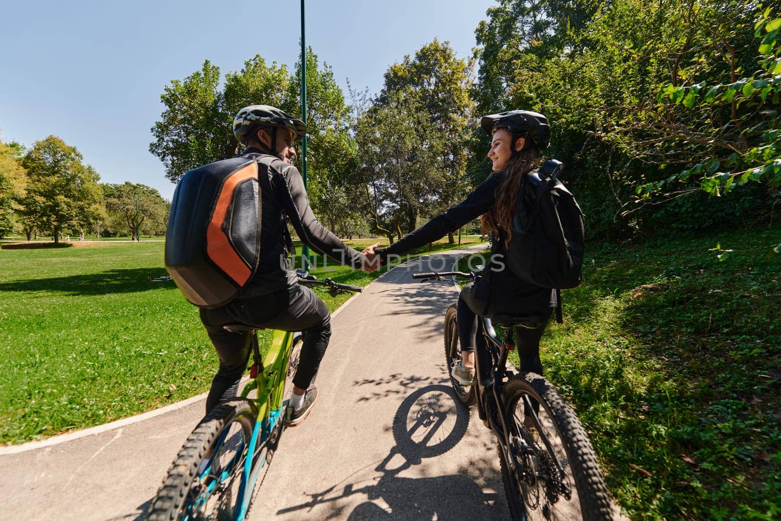 A sweet couple, adorned in cycling gear, rides their bicycles, their hands interlocked in a romantic embrace, capturing the essence of love, adventure, and joy on a sunlit path.