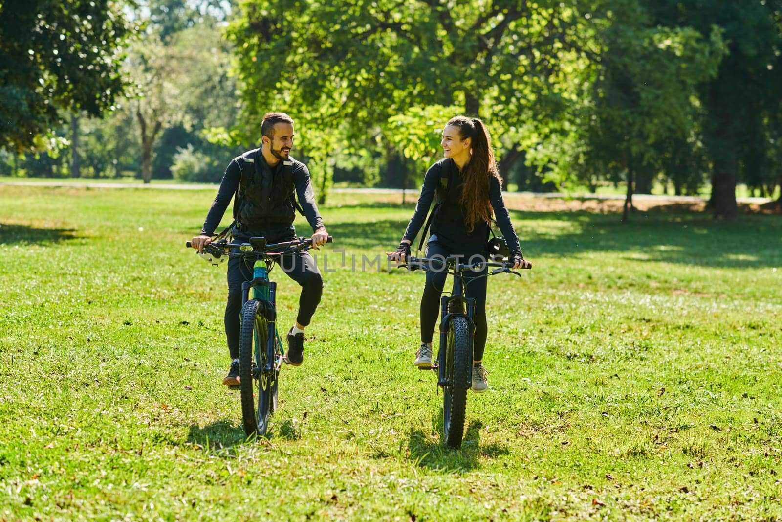 A blissful couple, adorned in professional cycling gear, enjoys a romantic bicycle ride through a park, surrounded by modern natural attractions, radiating love and happiness.