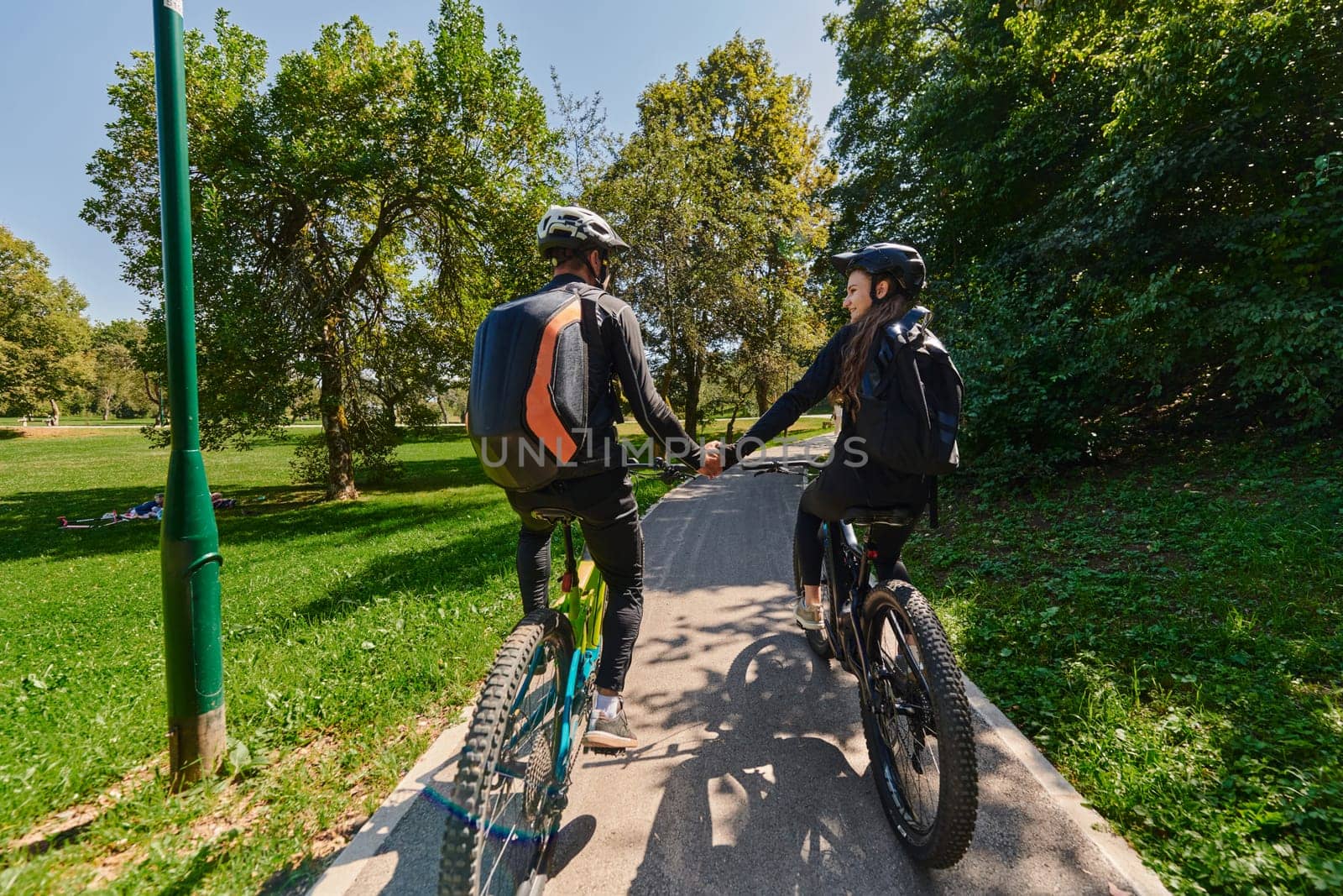 A sweet couple, adorned in cycling gear, rides their bicycles, their hands interlocked in a romantic embrace, capturing the essence of love, adventure, and joy on a sunlit path.