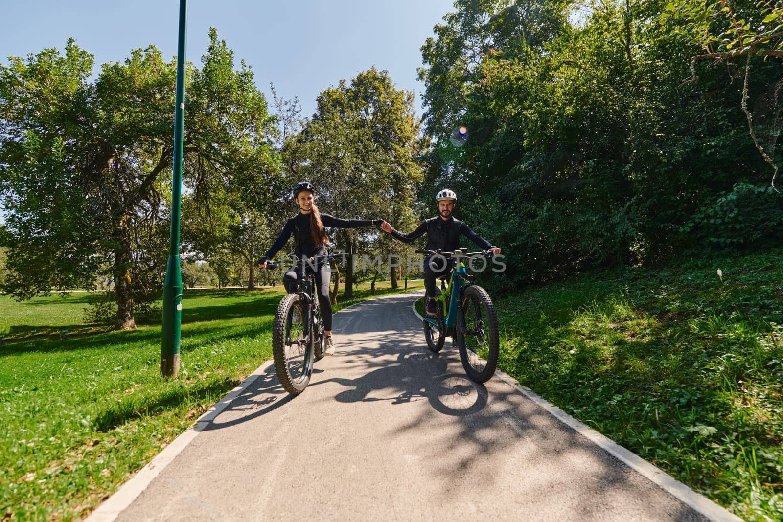 A sweet couple, adorned in cycling gear, rides their bicycles, their hands interlocked in a romantic embrace, capturing the essence of love, adventure, and joy on a sunlit path.