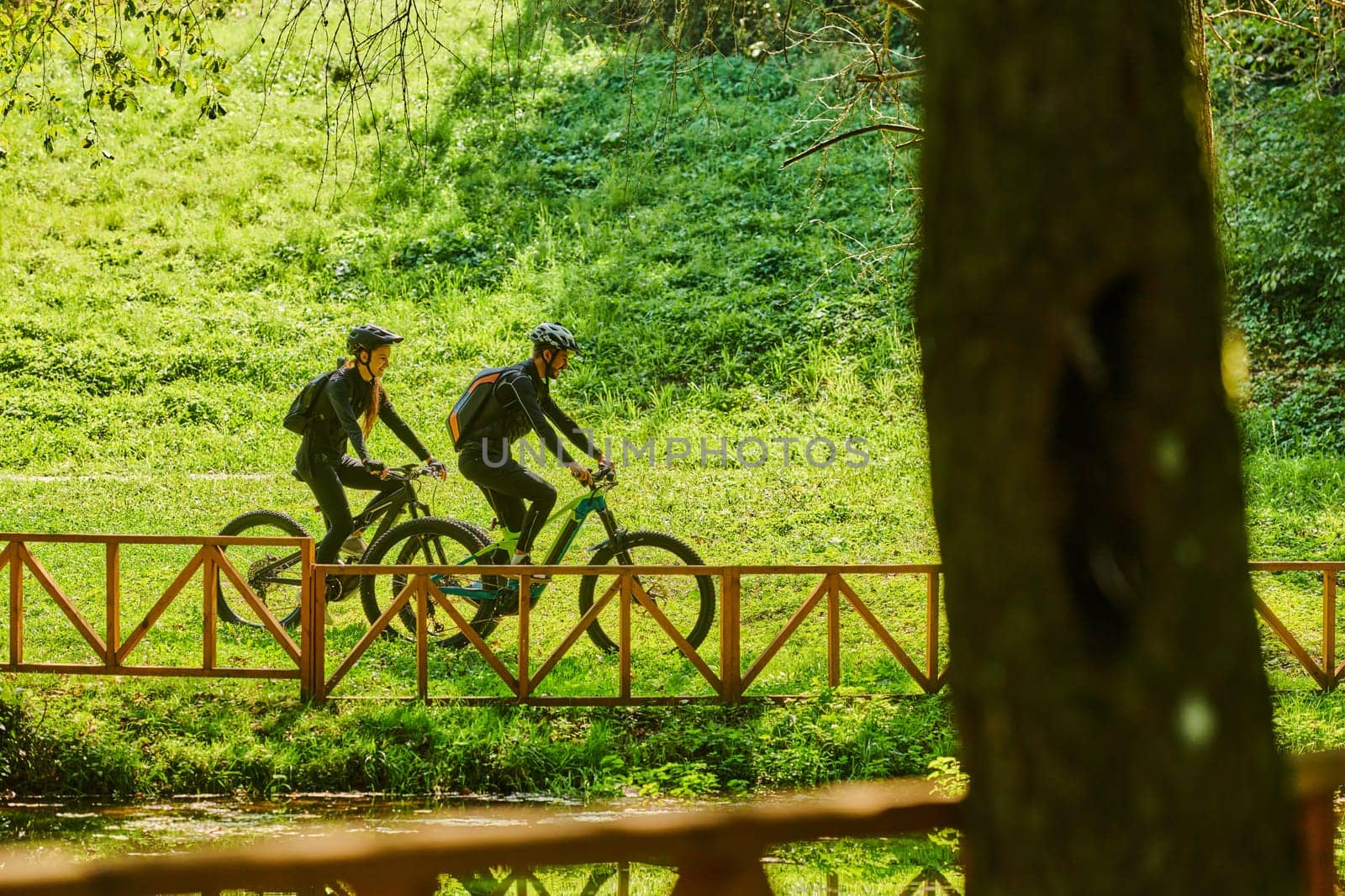 A blissful couple, adorned in professional cycling gear, enjoys a romantic bicycle ride through a park, surrounded by modern natural attractions, radiating love and happiness by dotshock