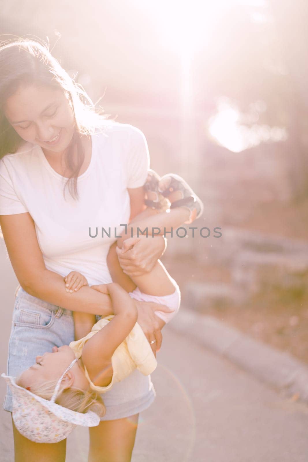 Smiling mom holding little girl upside down in bright sunlight by Nadtochiy