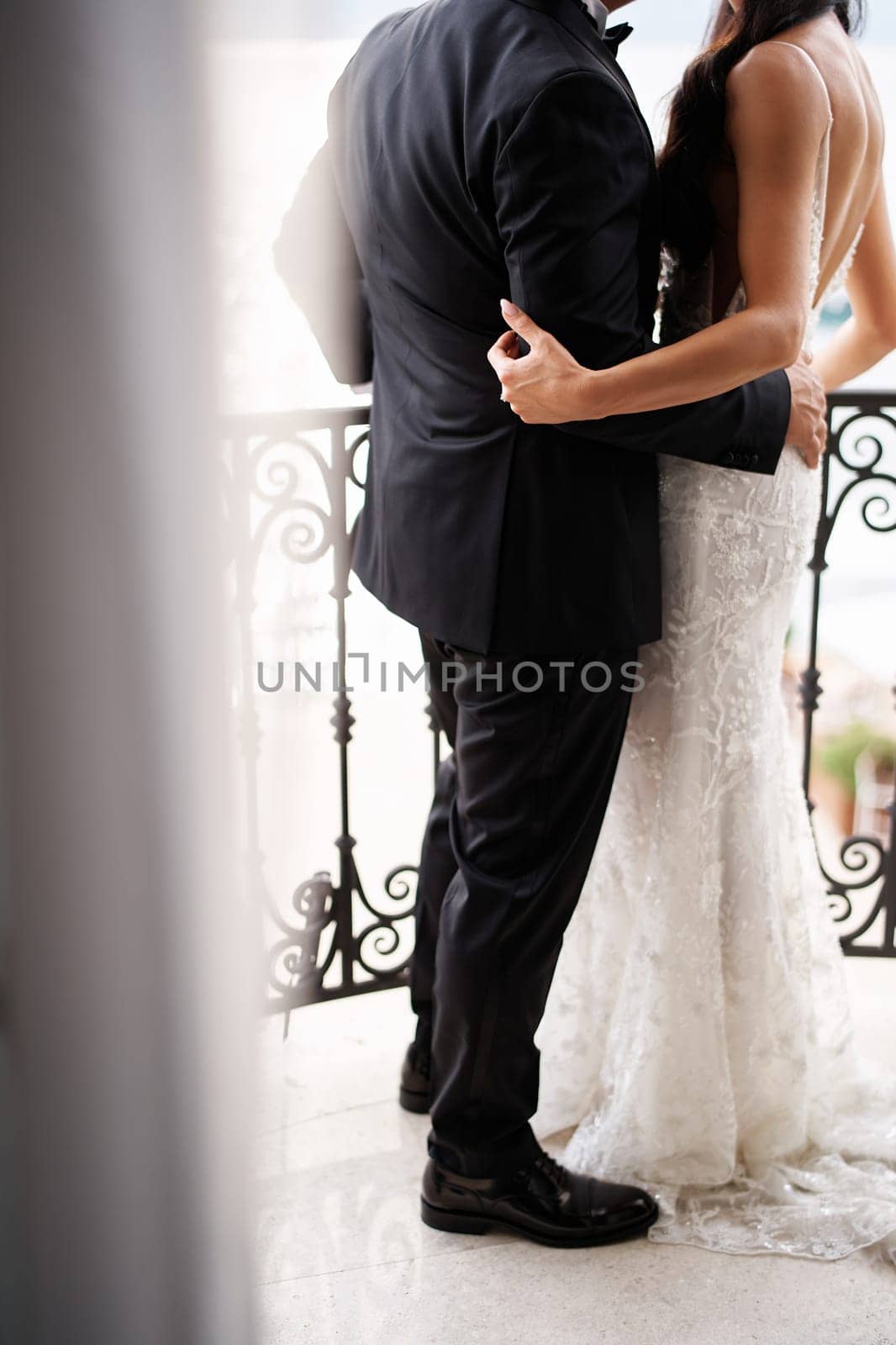 Groom hugs bride while standing on the balcony, looking at the sea. Back view. Cropped. Faceless. High quality photo
