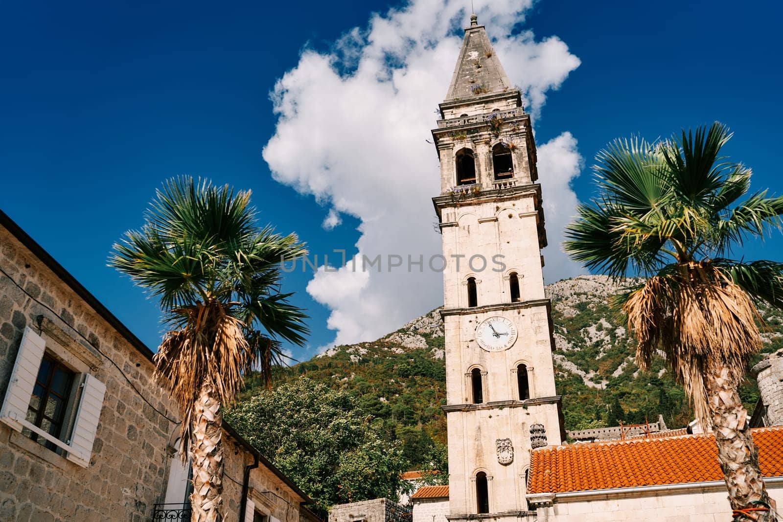 Bell tower of the Church of St. Nicholas with a clock among ancient houses against the backdrop of green mountains. Perast, Montenegro. High quality photo