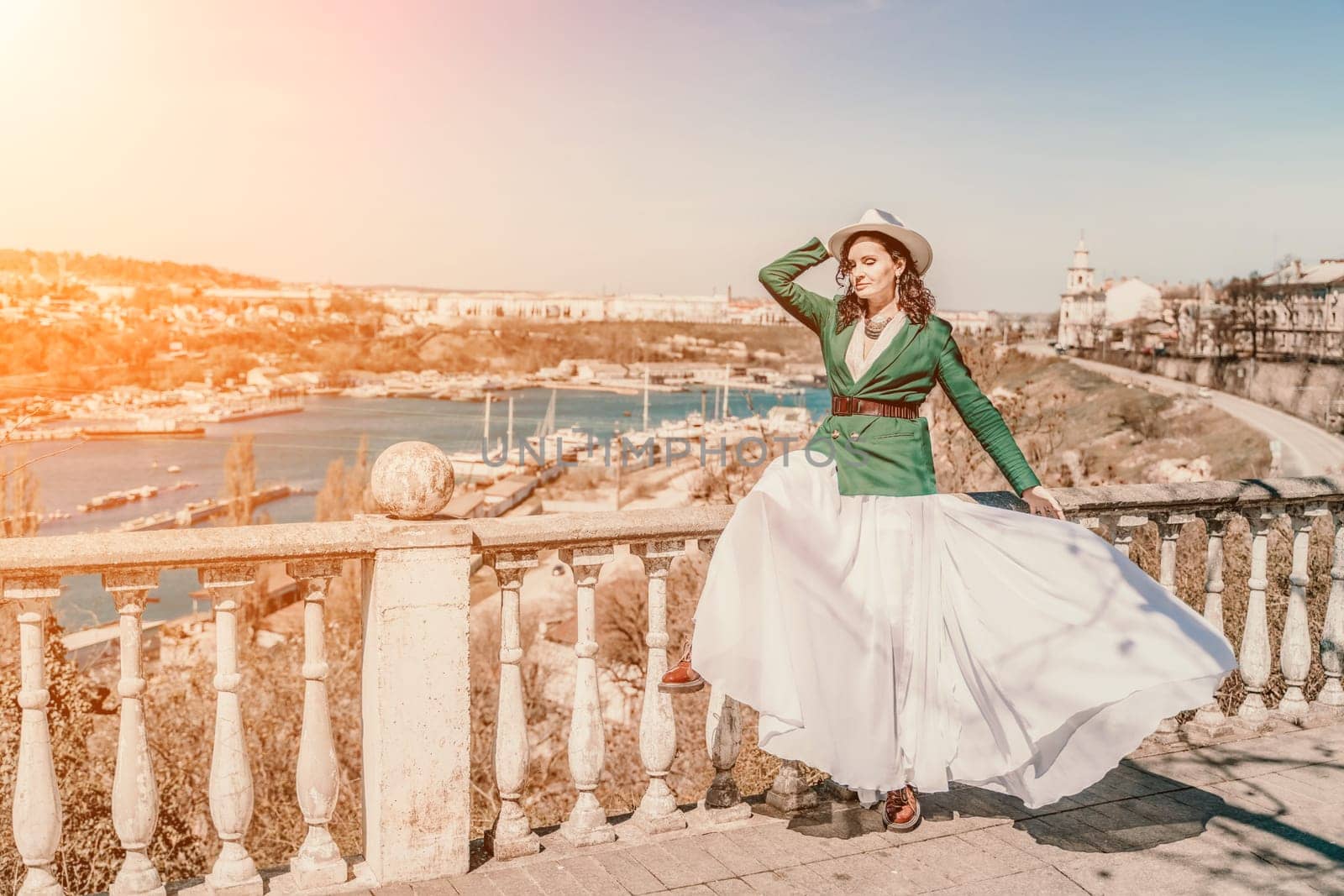 Woman walks around the city, lifestyle. A young beautiful woman in a green jacket, white skirt and hat is sitting on a white fence with balusters overlooking the sea bay and the city. by Matiunina