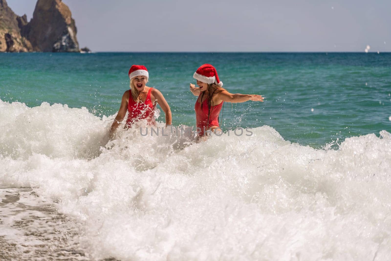 Women Santa hats ocean play. Seaside, beach daytime, enjoying beach fun. Two women in red swimsuits and Santa hats are enjoying themselves in the ocean waves