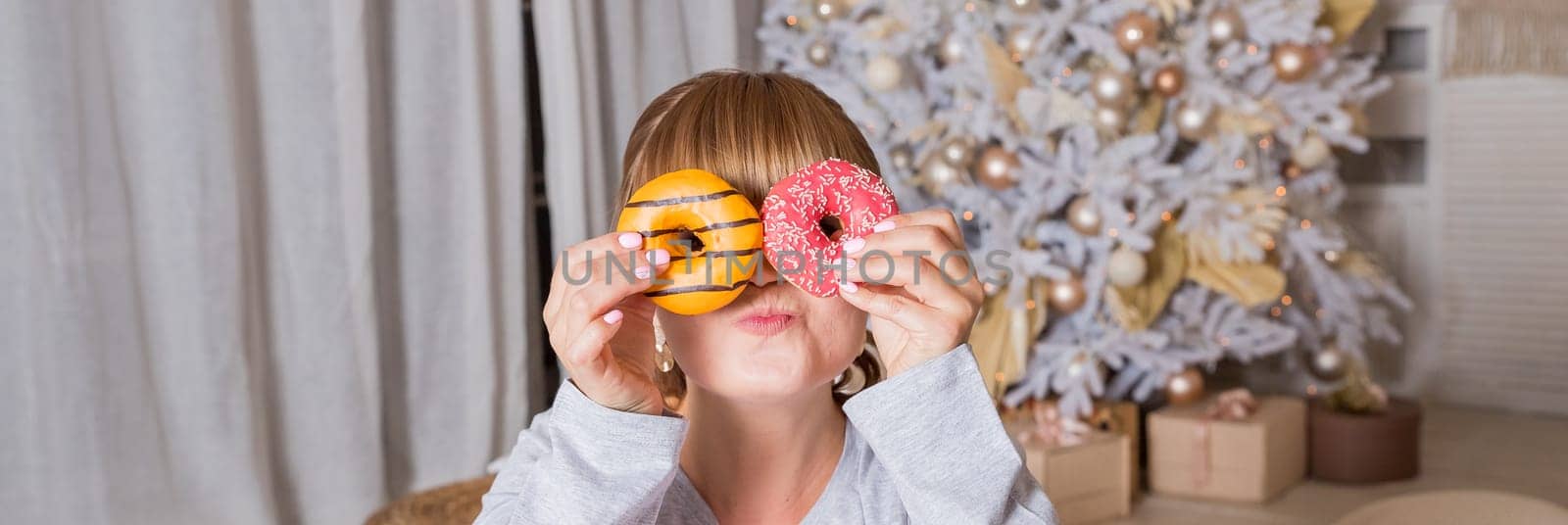 cheerful young woman holds donuts in her hands on christmas tree background, winter holidays. portrait of funny girl in pajamas covering her eyes with donuts by YuliaYaspe1979