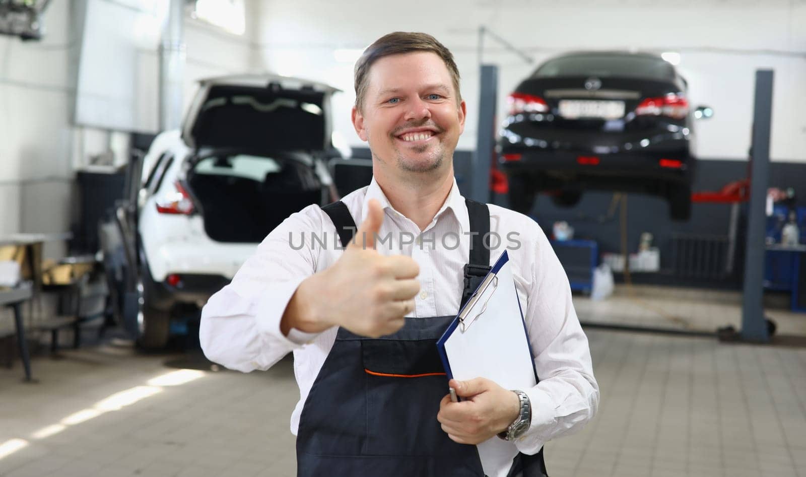 Portrait of young handsome man in uniform with thumbs up on background of car service. Recommendation quality car repair concept