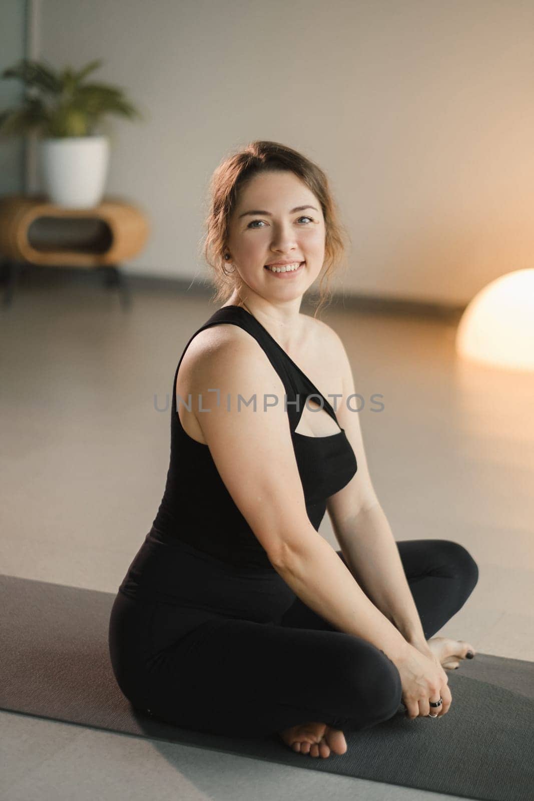 A girl in black sportswear is sitting on a yoga mat and smiling.
