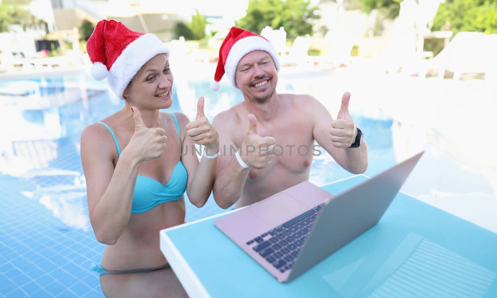 Family of bloggers in santa claus hats show thumbs up in laptop monitor standing in pool by kuprevich