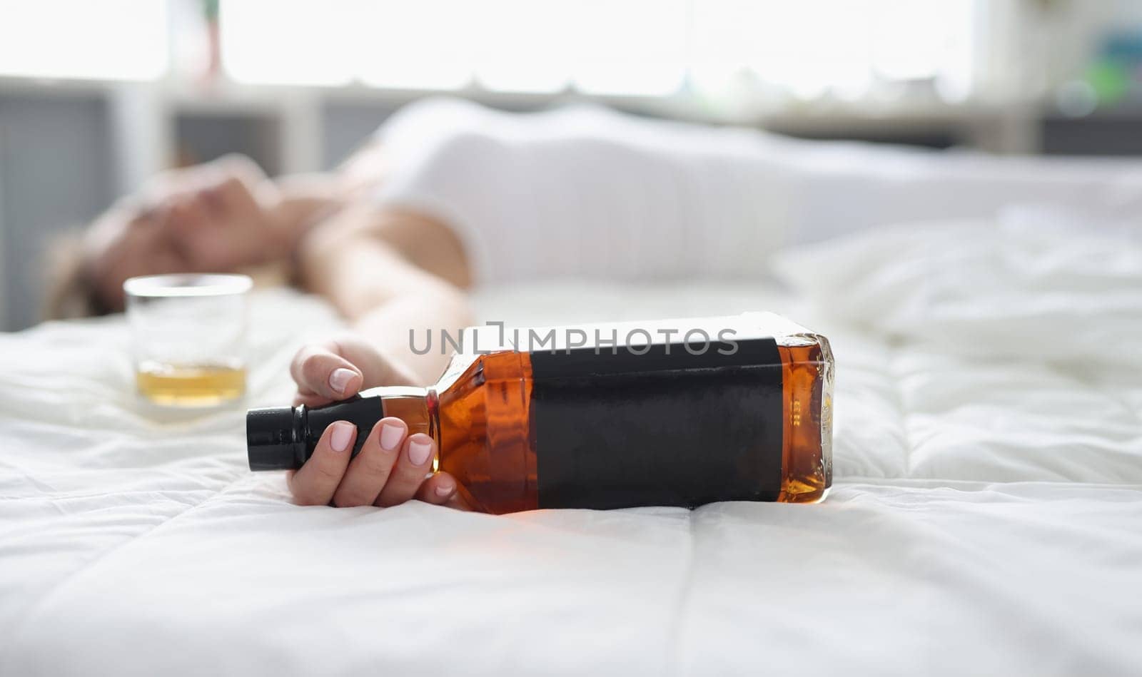 Woman lies in white bed with bottle of whiskey in hand. Alcohol addiction stress and depression concept