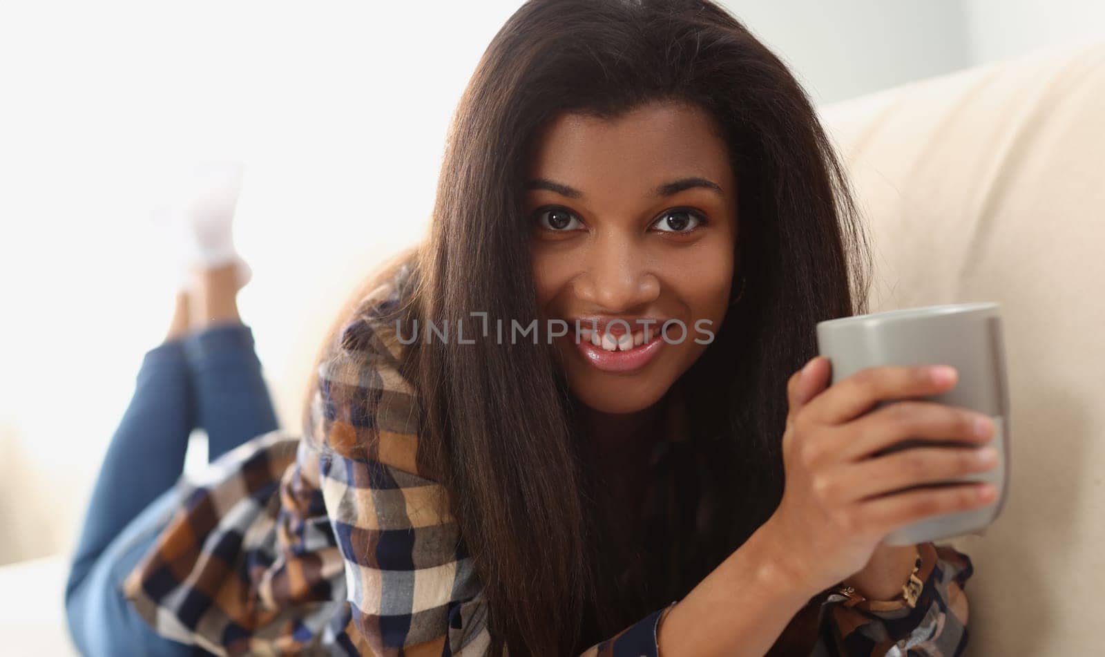 Young beautiful afro woman drinks coffee and lies on sofa. Home leisure and joyful mood concept