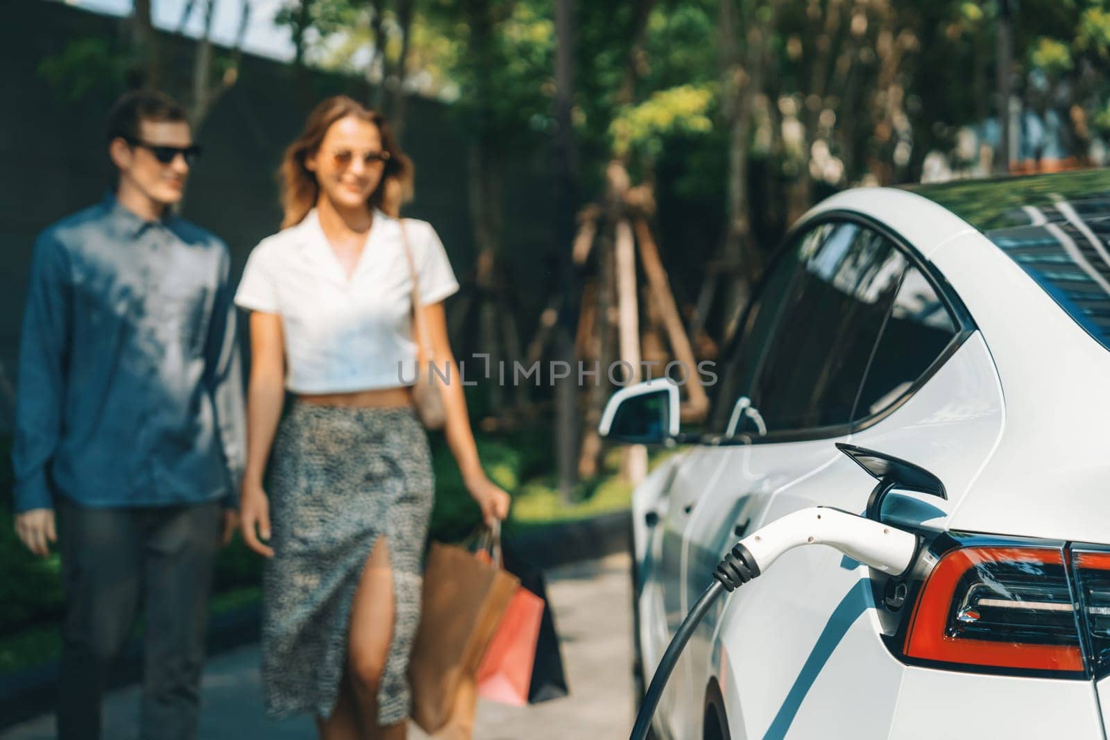 Young couple travel with EV electric car charging in green sustainable city outdoor garden in summer shows urban sustainability lifestyle by green clean rechargeable energy of electric vehicle innards