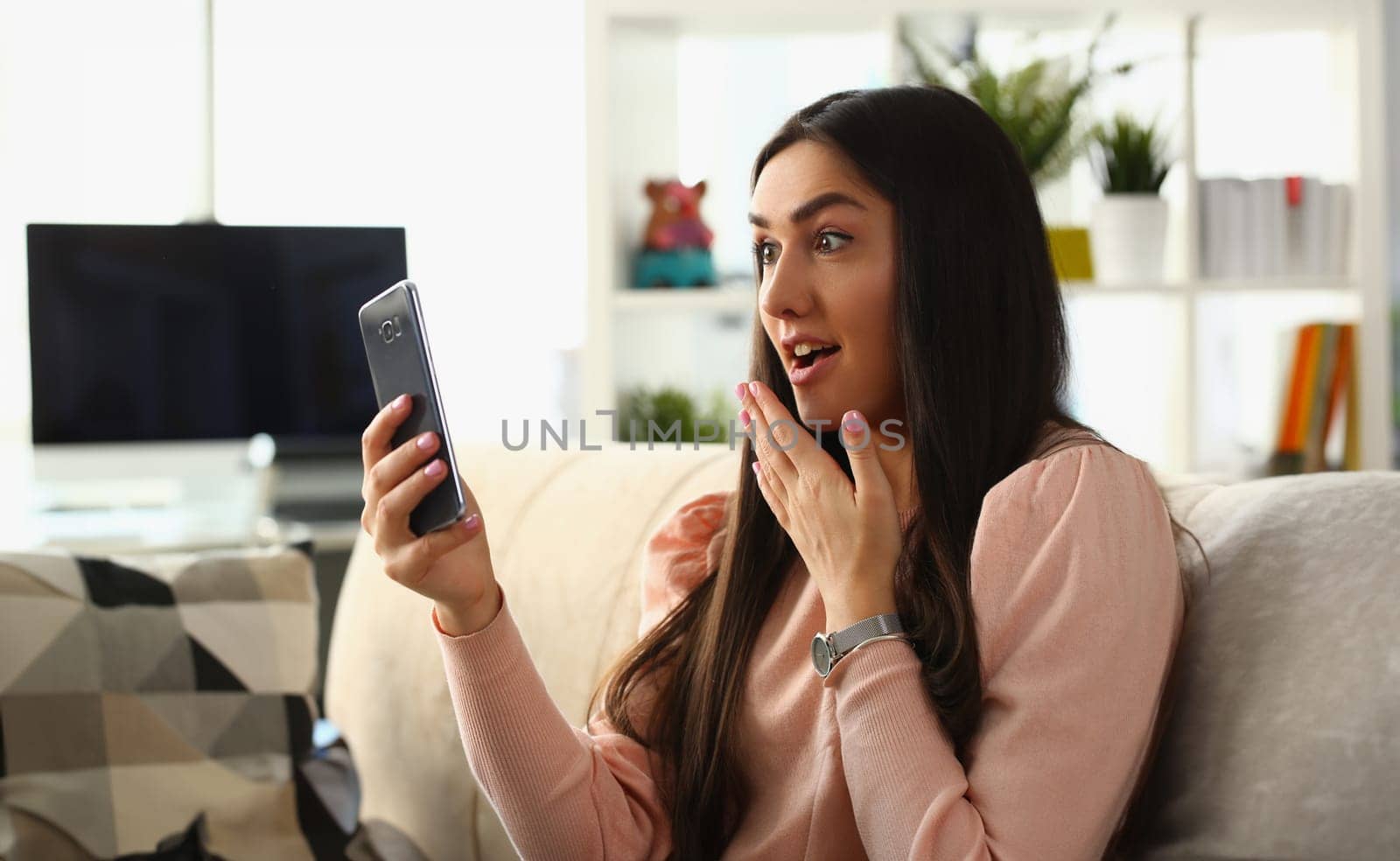 Woman surprised in shock looks at smartphone screen while sitting on couch at home. Emotion joy delight and good news concept