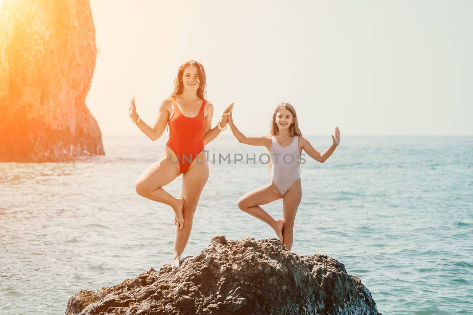 Woman and her daughter practicing balancing yoga pose on one leg up together on rock in the sea. Silhouette mother and daughter doing yoga at beach by panophotograph