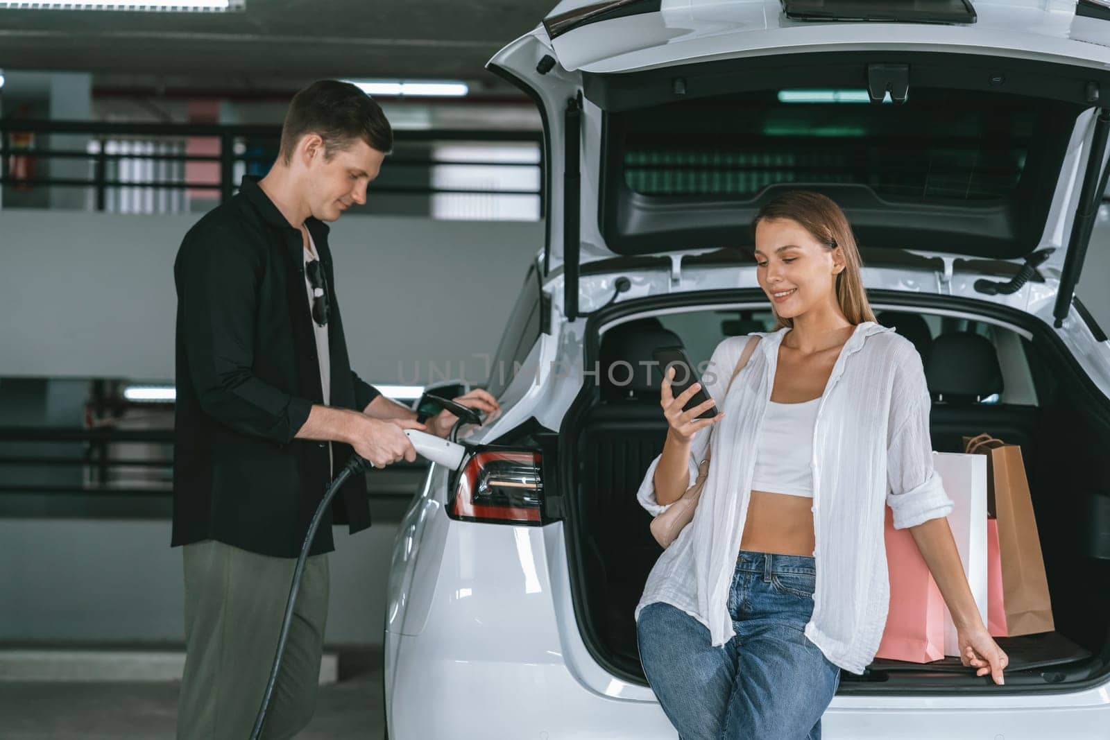 Young couple travel with EV electric car to shopping center parking lot charging in downtown city showing urban sustainability lifestyle by green clean rechargeable energy of electric vehicle innards