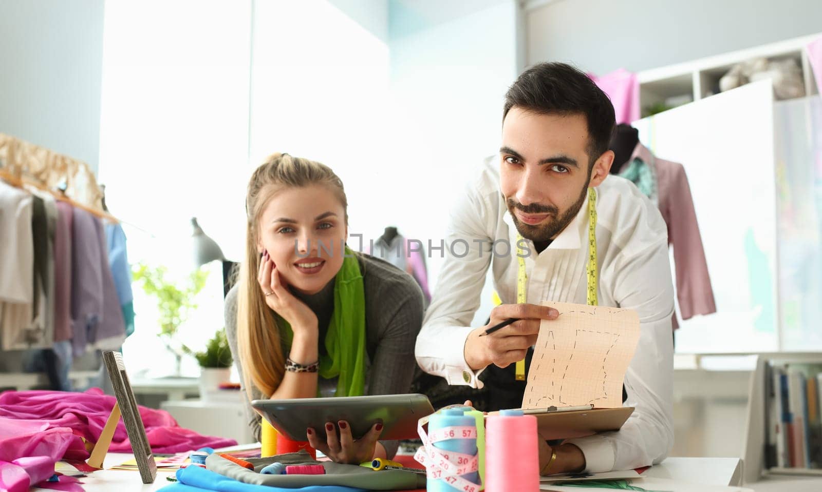 Young business woman stylist seamstress holding tablet male colleague showing sketches of clothes. Discussion of a new collection of clothes in creative sewing workshop