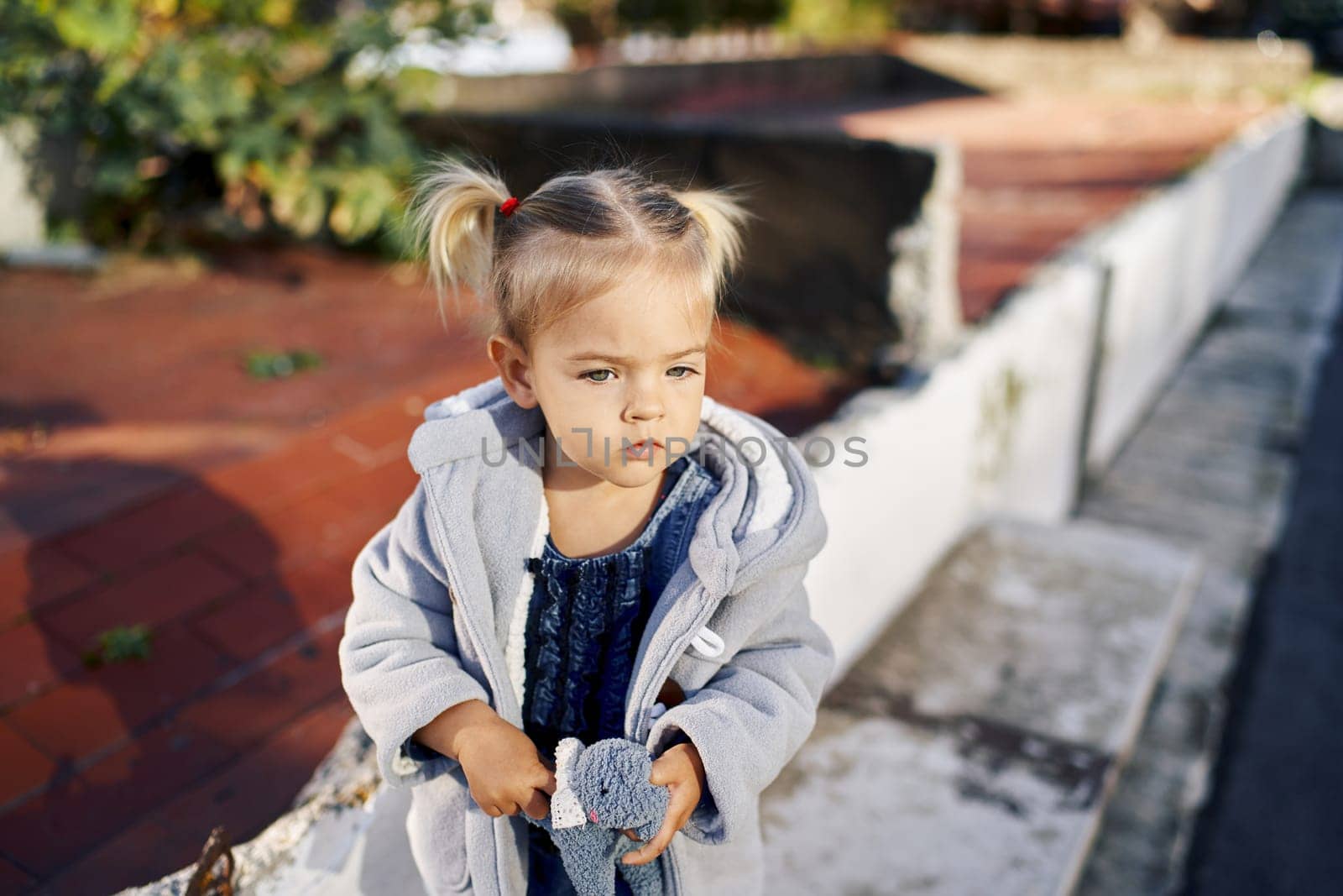 Little girl with a soft toy in her hands sits on a curb in the park. High quality photo
