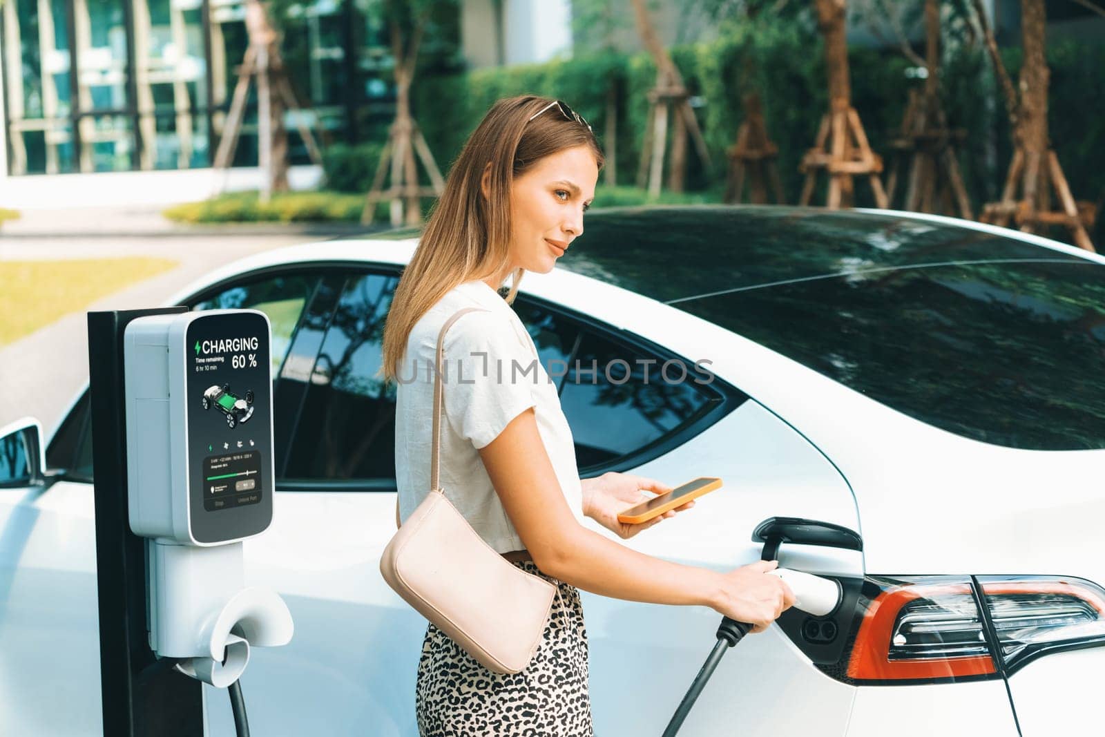 Young woman travel with EV electric car charging in green sustainable city outdoor garden in summer. Urban sustainability lifestyle by green clean rechargeable energy of electric BEV vehicle innards