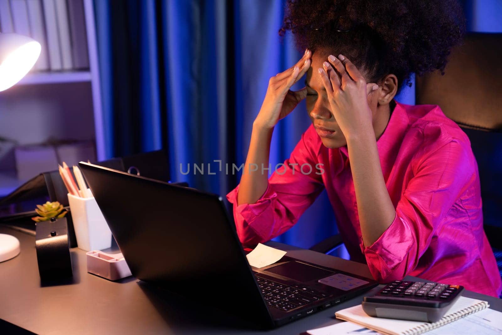 African woman businesswoman, wearing pink shirt having headache with migraine, sitting at computer laptop with leaning position. Concept of work form home with pressure and tension. Tastemaker.