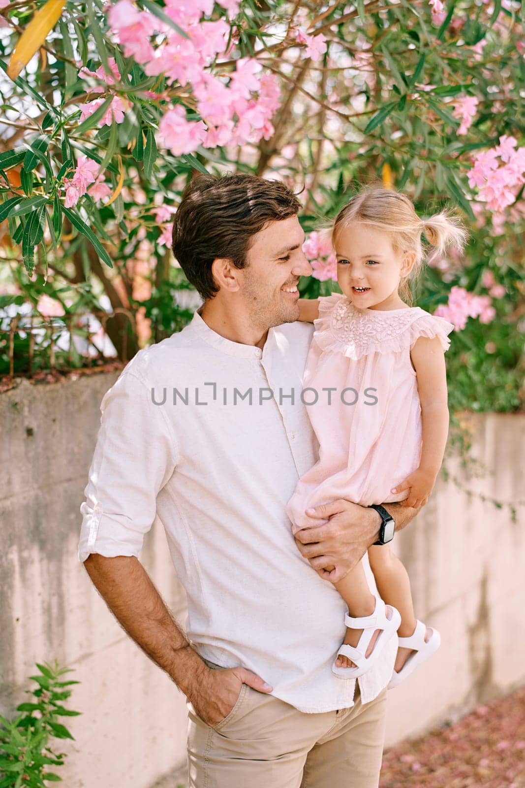 Smiling dad with a little girl in his arms stands near a flowering tree in the garden by Nadtochiy