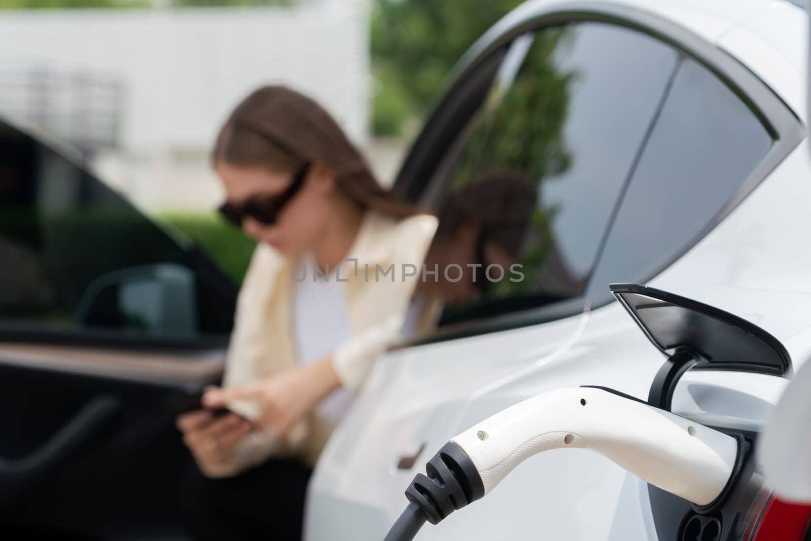 Young woman use smartphone to pay for electricity at public EV car charging station green city park. Modern environmental and sustainable urban lifestyle with EV vehicle. Expedient