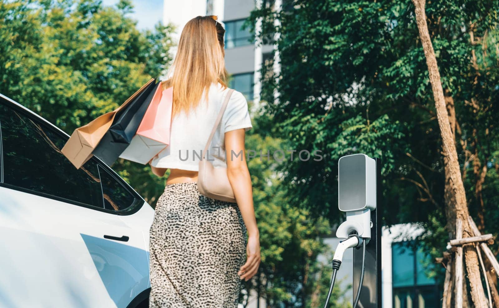 Young woman travel with EV electric car charging in green sustainable city outdoor garden in summer. Urban sustainability lifestyle by green clean rechargeable energy of electric BEV vehicle innards