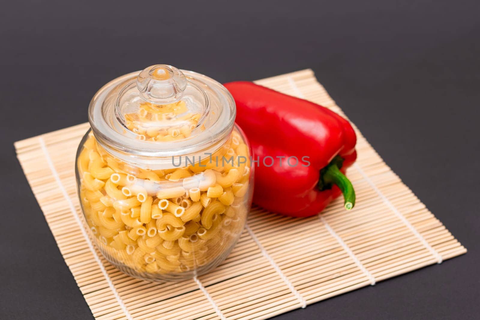 Uncooked Chifferi Rigati Pasta in Glass Jar with Red Bell Pepper on Bamboo Mat on Black Background. Fat and Unhealthy Food. Scattered Classic Dry Macaroni. Italian Culture and Cuisine. Raw Pasta