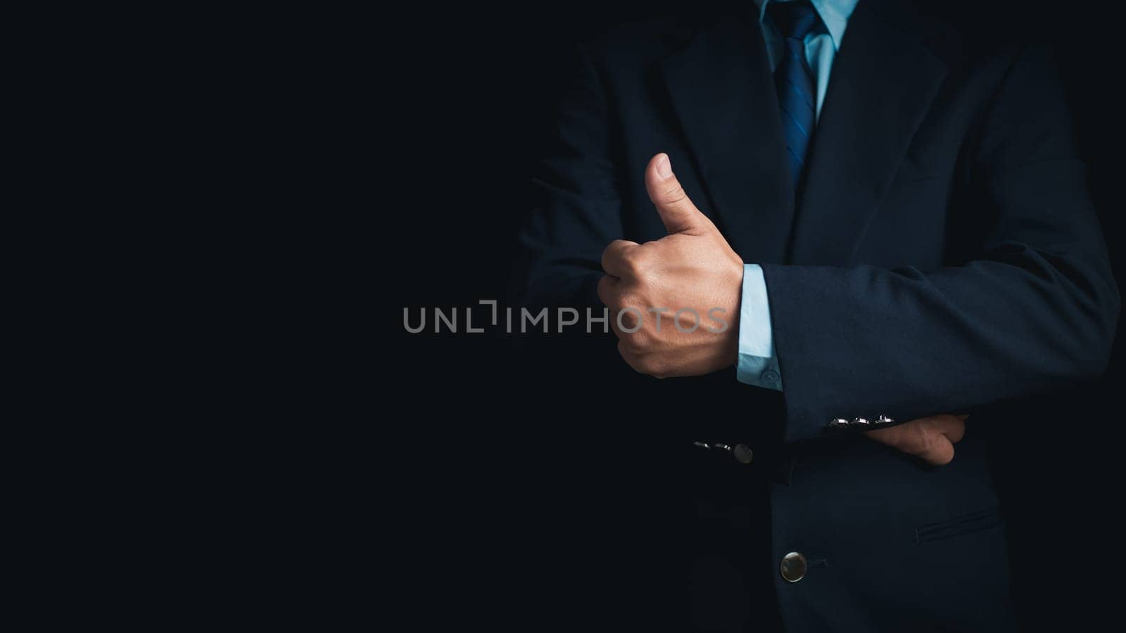 A businessman in a suit stands with his arms crossed and thumbs up on a dark background. by Unimages2527