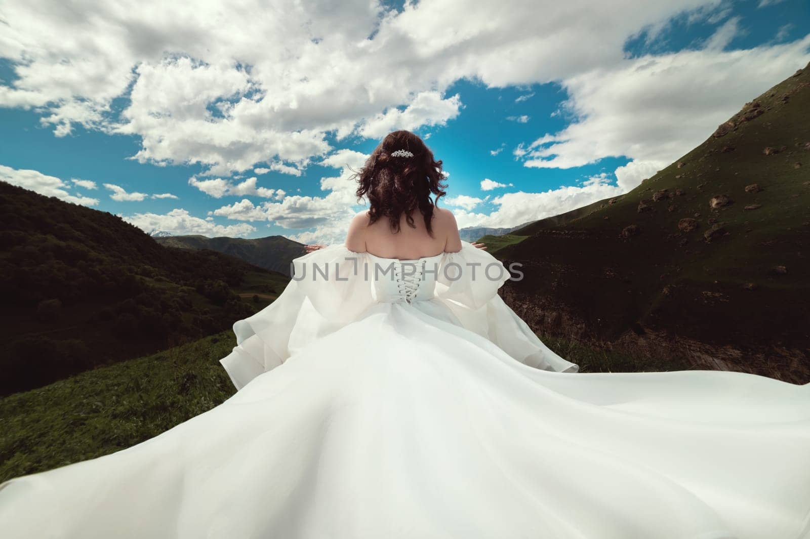 A girl in a white dress with a long flying train runs across a field in the green mountains on a sunny day