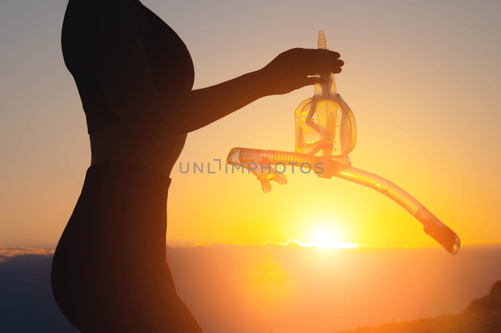 A hand holds snorkeling goggles (diving mask) against the background of the sunset sky. Silhouette of a sexy unrecognizable woman holding scuba gear in her hands