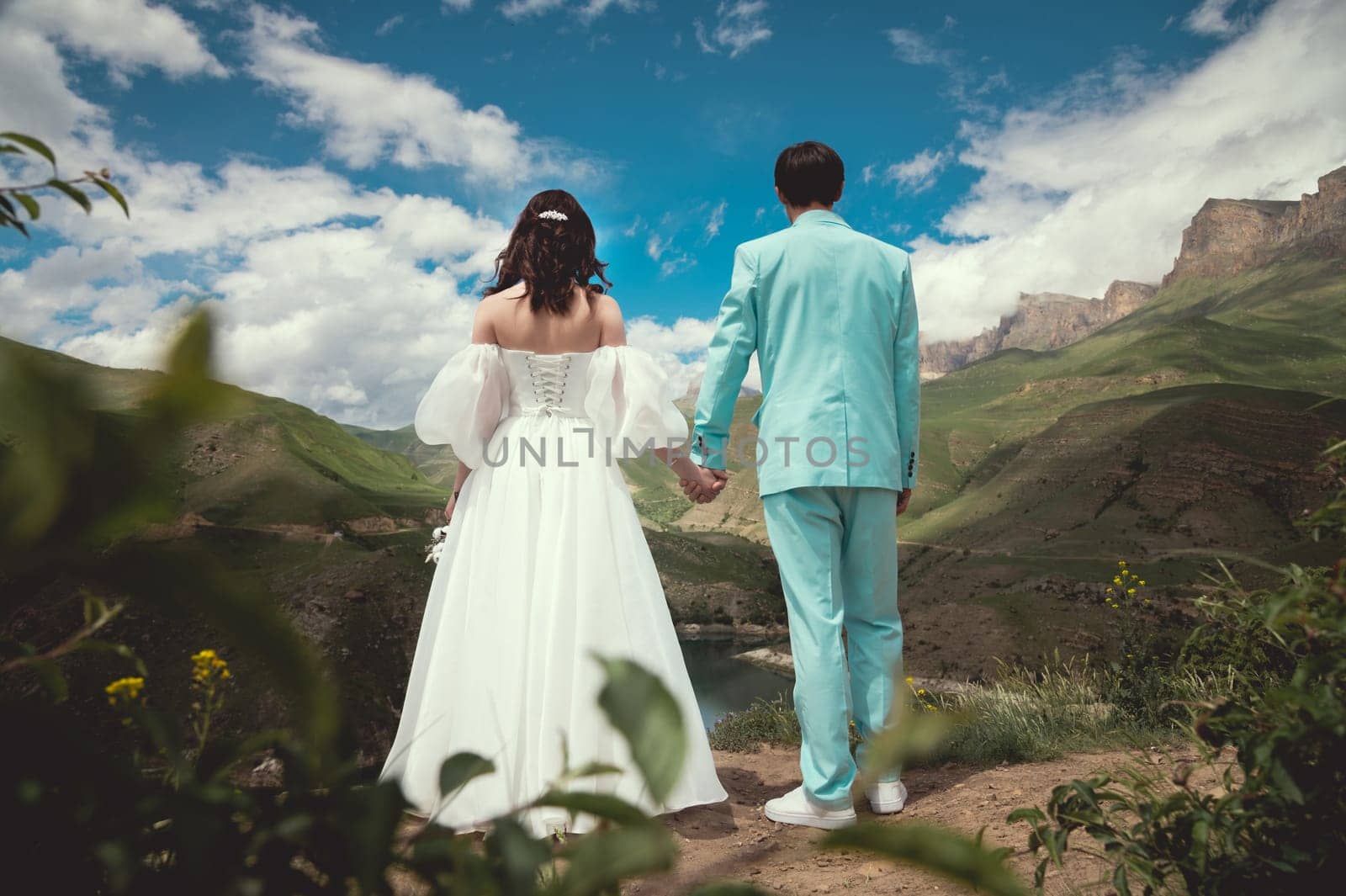Beautiful newlyweds stand sideways to each other, holding hands against the backdrop of sunset mountains. Romantic honeymoon.