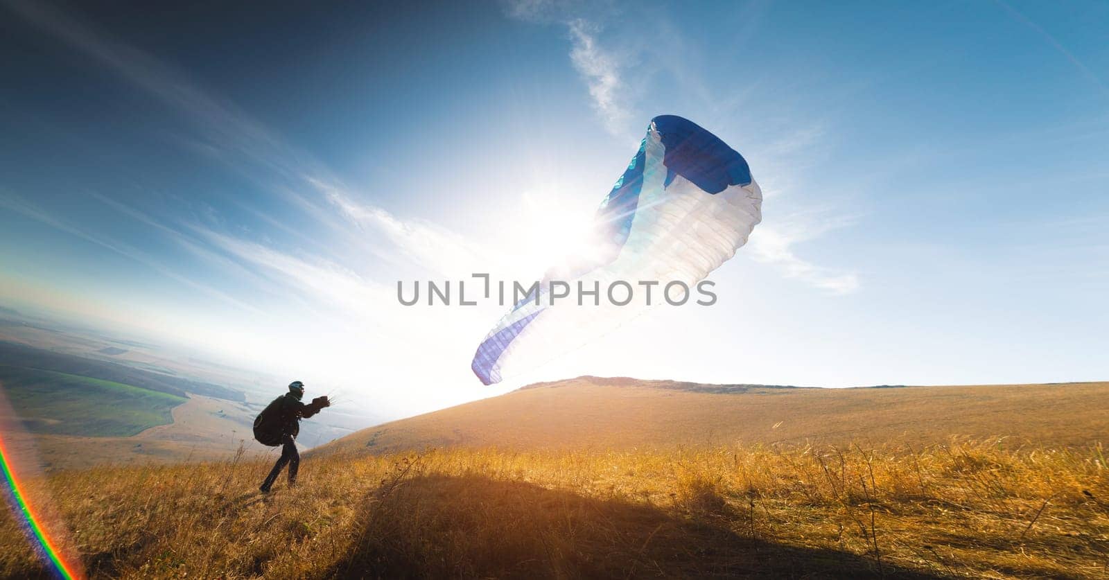 A paraglider with a blue parachute takes off. A man takes off and lands on a yellow field. a man preparing to take off with an open paraglider already in the air