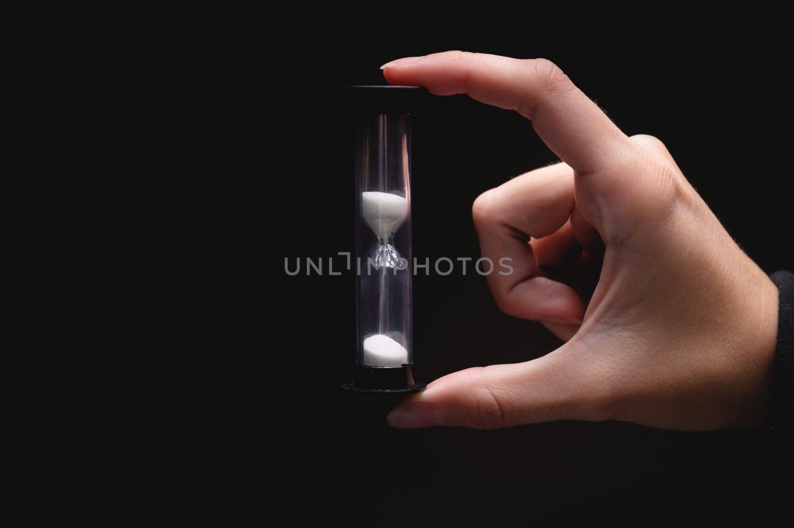 Close-up of a woman holding an hourglass in the dark. Fingers hold on both sides a plastic sand clock with white sand on a black background by yanik88