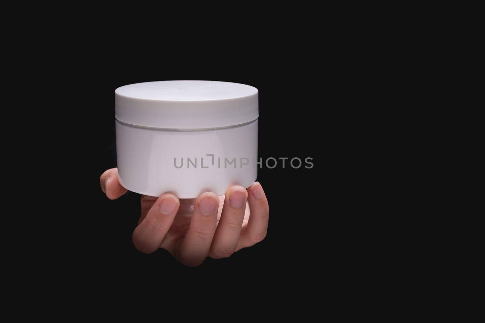 White cosmetic packaging mockup. A woman's hand holds a round jar of body cream on a black background. Beauty industry concept. Close-up, copy space, banner.