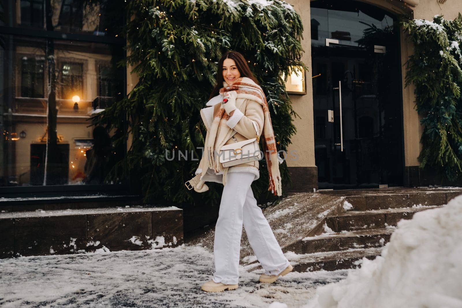 A girl with long hair in a scarf and with a white handbag walks down the street in winter by Lobachad