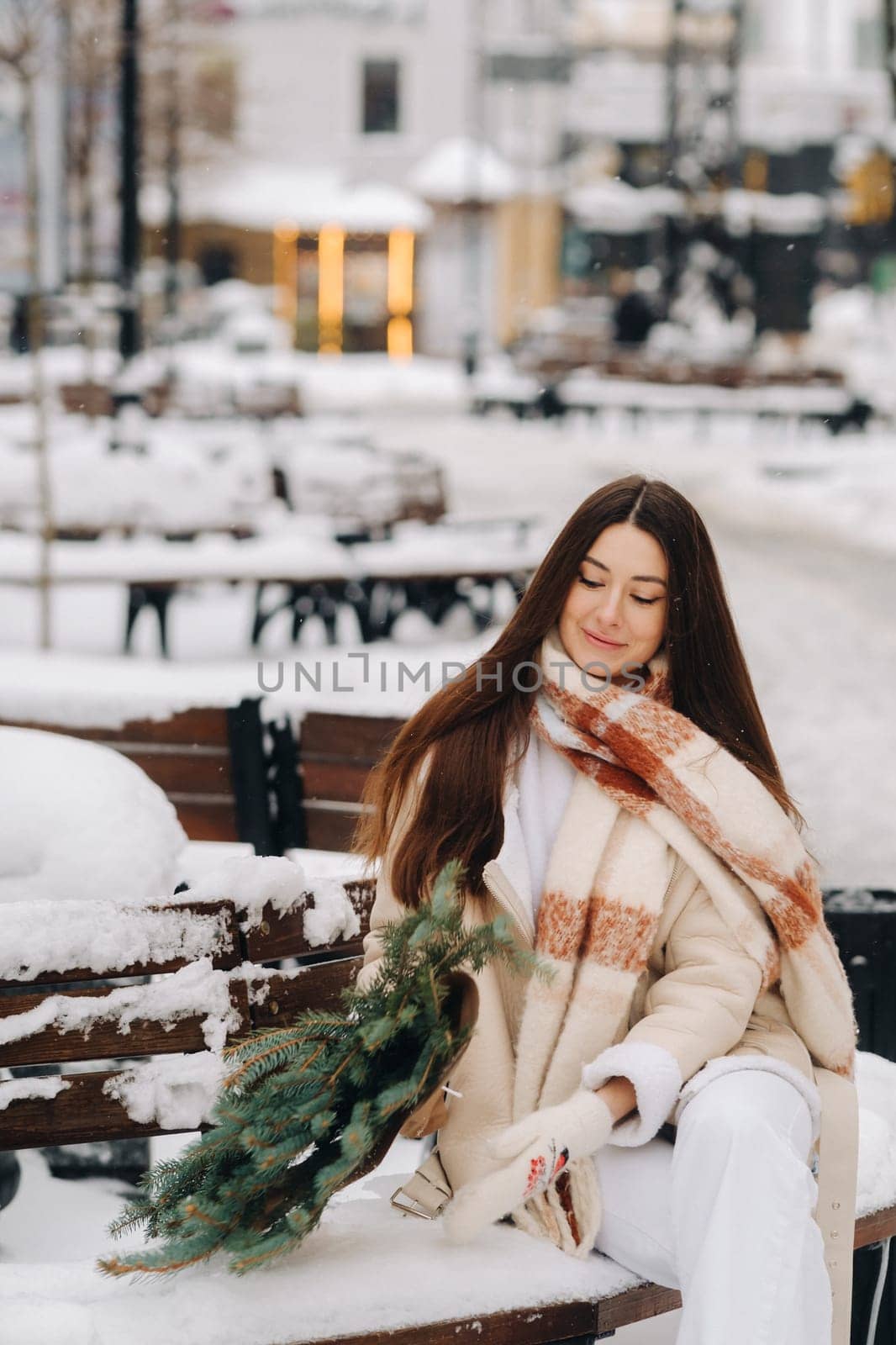 A girl with long hair in winter sits on a bench outside with a bouquet of fresh fir branches.